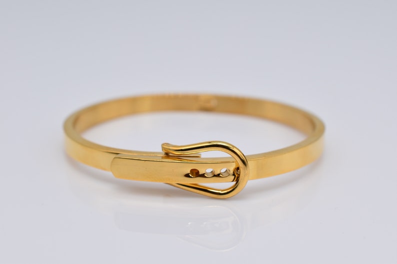 Op de afbeelding: Een gouden armband met een gespsluiting. De gesp heeft drie kleine, heldere stenen.