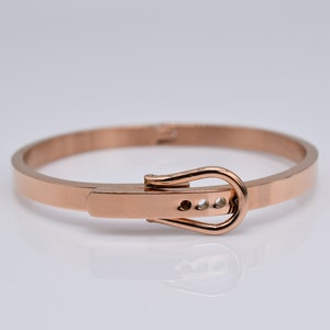 Op de afbeelding: Een armband van ros&eacute;goudkleurig metaal met een gespsluiting. De gesp heeft een kleine rij heldere stenen.