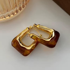 May include: A pair of gold-toned, square hoop earrings with a tortoiseshell pattern. The earrings have a unique, textured gold frame and a warm, amber-brown center, creating a stylish and eye-catching accessory.