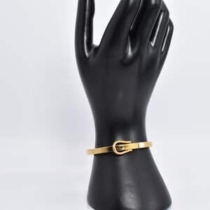 Op de afbeelding: Een gouden metalen armband met een gespsluiting. De armband is op een zwart handmodel.
