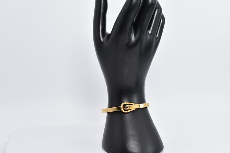 Op de afbeelding: Een gouden metalen armband met een gespsluiting. De armband is op een zwart handmodel.