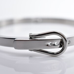 Op de afbeelding: Een zilveren metalen armband met een gespsluiting. De gesp heeft twee kleine, heldere stenen.