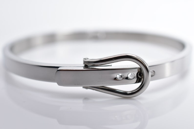 Op de afbeelding: Een zilveren metalen armband met een gespsluiting. De gesp heeft twee kleine, heldere stenen.