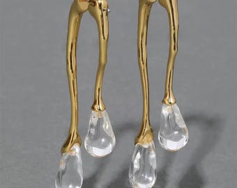 May include: A pair of gold-toned earrings featuring a long, curved, organic metal stem that splits into two branches. Each branch terminates in a clear, teardrop-shaped resin pendant that hangs at different lengths, creating a fluid, asymmetrical look.