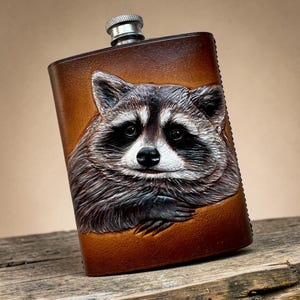 Op de afbeelding: Een met bruin leer beklede metalen heupfles met een gedetailleerd, in reliëf gemaakt en handgeschilderd portret van een wasbeer op de voorzijde. De fles heeft een zilverkleurige schroefdop en een afgeronde rechthoekige vorm met zichtbare stiksels aan de zijkant.