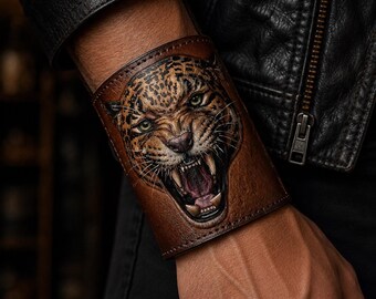 May include: A wide brown leather cuff bracelet featuring a detailed, embossed, and hand-painted illustration of a snarling leopard with bared teeth, whiskers, and intense green eyes, finished with dark stitched edges along the top and bottom.