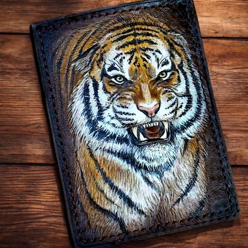 May include: A handcrafted leather wallet featuring a detailed, embossed tiger face. The tiger's fur is rendered in shades of brown, tan, and white, with black stripes. The wallet has a dark brown border and a stitched edge, showcasing a unique animal design.