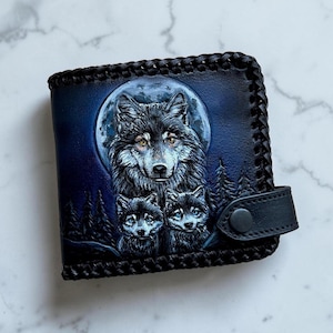 Pode incluir: Uma carteira de couro preta com uma cena esculpida e pintada à mão de um lobo grande e dois filhotes diante de uma lua cheia e uma floresta. A carteira possui uma borda de couro trançado e uma tira de couro preta com fecho de botão de pressão.