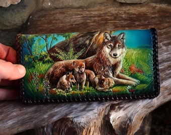 May include: A rectangular leather wallet featuring a hand-painted scene of a large wolf with three pups in a lush green forest setting. The wallet has a dark braided leather border and displays vibrant shades of brown, green, and blue.
