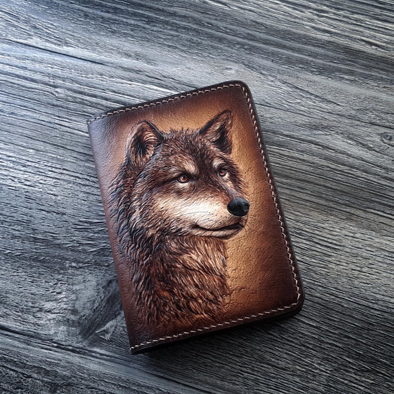 May include: A brown leather wallet with a detailed carving of a wolf's face. The wolf is looking to the right and has a light brown fur.