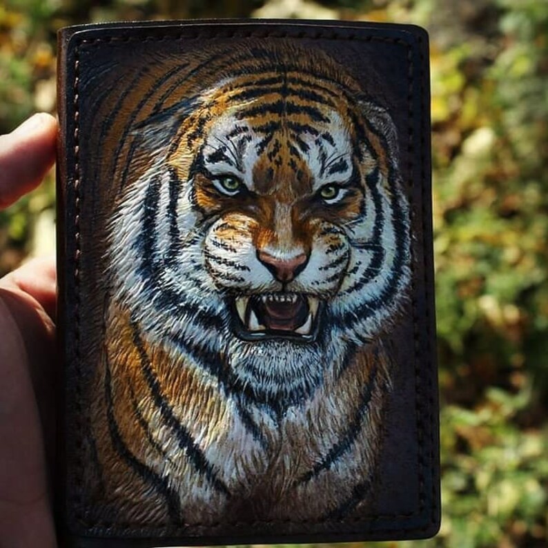 May include: Brown leather wallet featuring a detailed, hand-painted tiger face. The tiger has orange and black stripes, white fur, and green eyes. The wallet is held in a hand, with a blurred green background.