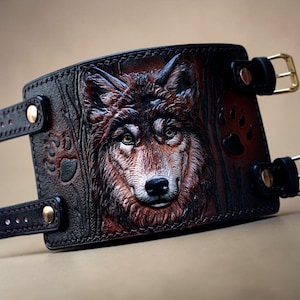 May include: A brown leather wrist cuff featuring a detailed relief carving of a wolf's face. The cuff has adjustable straps with gold-colored buckles and is decorated with paw prints. The leather is a rich, dark brown with lighter highlights.