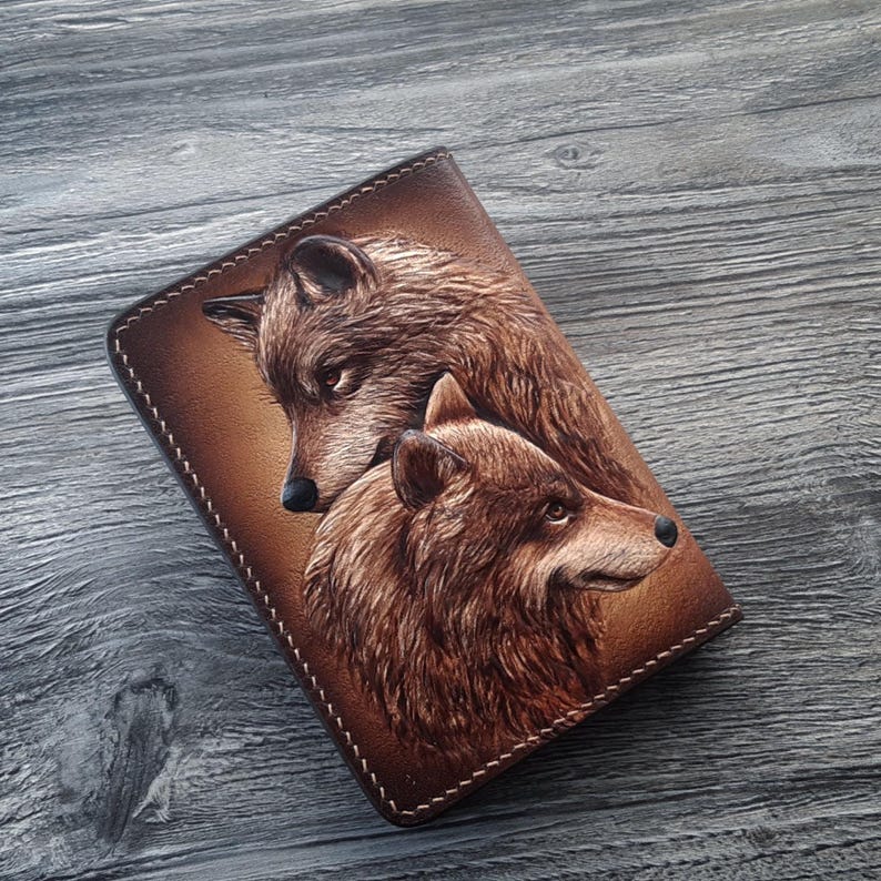 May include: A brown leather notebook cover with a tooled design of two wolves. The wolves are facing each other, with one wolf's head turned slightly to the side. The cover has a stitched edge.
