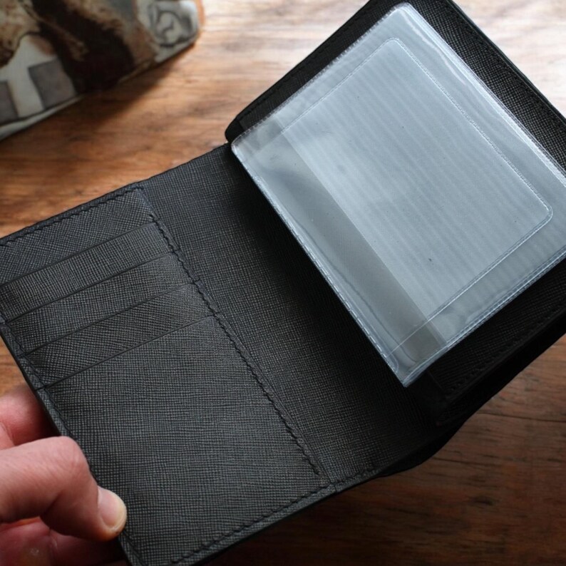May include: Open black wallet with multiple card slots and a clear plastic ID holder. The wallet is made of textured black material and is open, displaying the interior design. The background is a wooden surface.