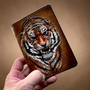 Pode incluir: Uma carteira de couro marrom com um rosto de tigre em relevo detalhado. O tigre tem listras laranja e pretas, pelo branco e dentes visíveis. A carteira é retangular com uma borda costurada.
