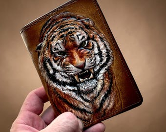 May include: A brown leather wallet featuring a detailed, embossed tiger face. The tiger has orange and black stripes, white fur, and visible teeth. The wallet is rectangular with a stitched border.