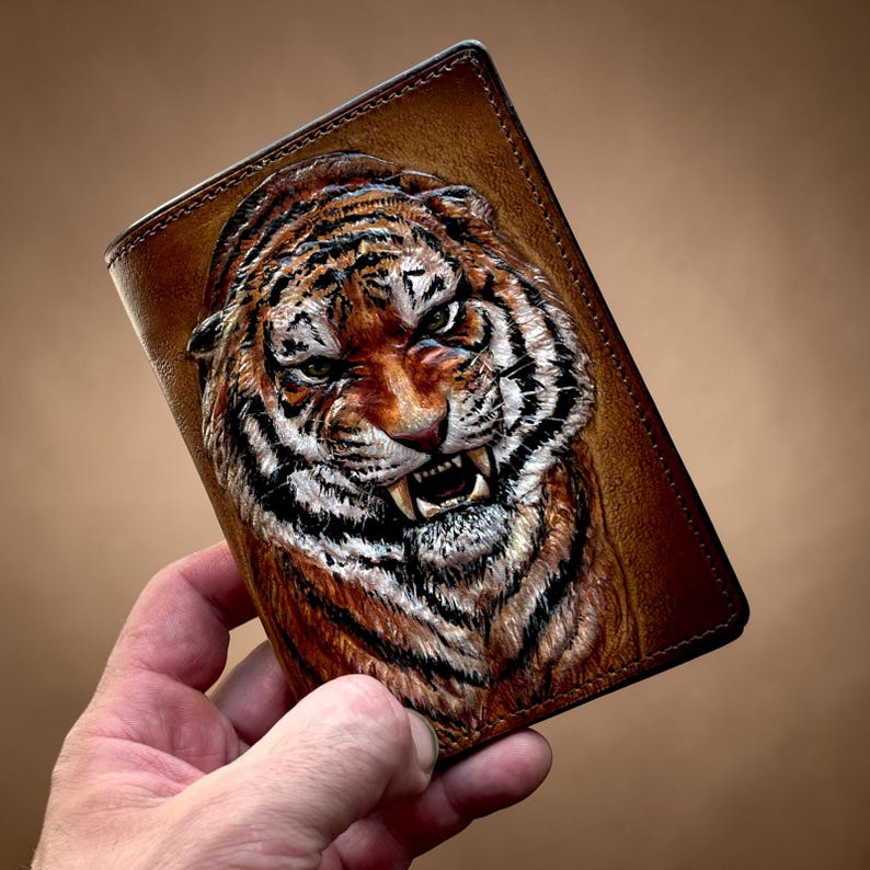 May include: A brown leather wallet featuring a detailed, embossed tiger face. The tiger has orange and black stripes, white fur, and visible teeth. The wallet is rectangular with a stitched border.
