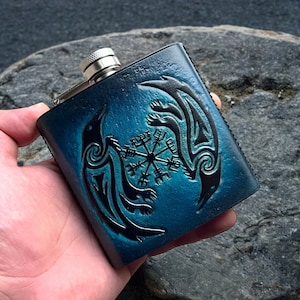 May include: A blue leather flask with a black design of two ravens and a compass. The flask has a silver cap.