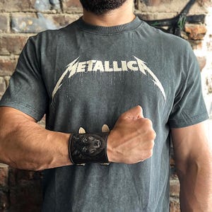Può includere: Una t-shirt grigio scuro a maniche corte con il logo bianco Metallica sul petto. Un ampio bracciale a fascia in pelle nera è indossato al polso, mostrando un ritratto dettagliato in rilievo di un cane con inserti in pelle chiara.