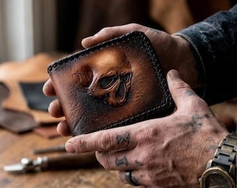 May include: A brown leather bifold wallet featuring a three-dimensional embossed skull design on the front surface. The wallet has dark, burnished edges and visible stitching along the perimeter, showcasing a textured, weathered finish.
