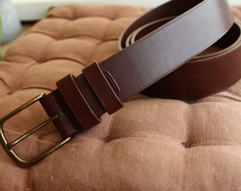 May include: A brown leather belt with a gold metal buckle. The belt is folded in half and lying on a brown fabric surface.
