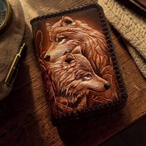May include: A rectangular brown leather wallet featuring a detailed embossed design of two wolves. The leather surface displays intricate fur textures and facial features of the animals. The edges of the wallet are finished with a dark braided leather trim.