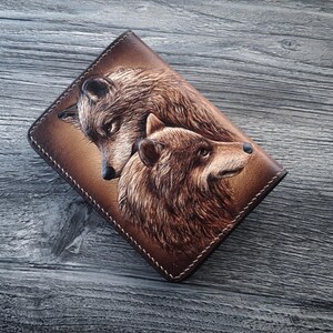 May include: A brown leather wallet with a tooled design of two wolves facing each other. The wolves are brown and have detailed fur.