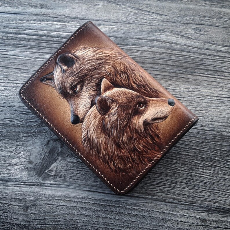 May include: A brown leather wallet with a tooled design of two wolves facing each other. The wolves are brown and have detailed fur.