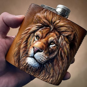 Puede incluir: Una petaca de cuero marrón con una imagen en relieve detallada de la cara de un león. La petaca tiene una tapa plateada y forma rectangular. La melena y la cara del león están intrincadamente talladas, mostrando tonos marrones y beige.