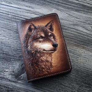 May include: A brown leather notebook cover with a detailed embossed image of a wolf's head. The wolf's fur is depicted in shades of brown and white, and its eyes are a piercing blue.