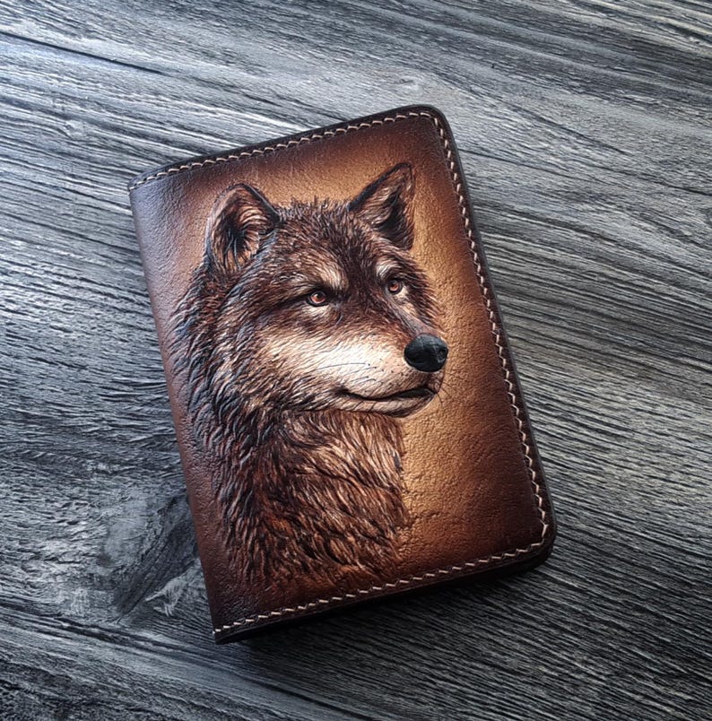May include: A brown leather notebook cover with a detailed embossed image of a wolf's head. The wolf's fur is depicted in shades of brown and white, and its eyes are a piercing blue.