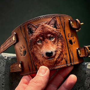 May include: A brown leather cuff bracelet featuring a detailed relief of a wolf's face. The cuff has a buckle closure and decorative paw prints. The wolf has green eyes and is surrounded by stylized tree branches.
