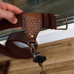 Op de afbeelding: Een bruine leren riem met een geweven ontwerp en een sleutelhanger eraan. De sleutelhanger heeft een metalen clip en een set sleutels.
