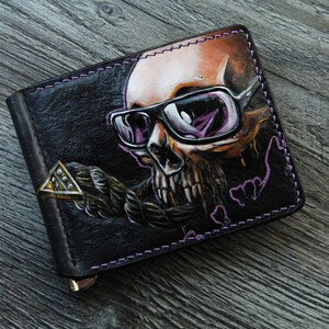 Könnte beinhalten: Eine schwarze Ledergeldbörse mit einem Totenkopfdesign. Der Totenkopf trägt eine Sonnenbrille und hat ein Seil um den Hals. Die Geldbörse hat einen Metallverschluss und lilafarbene Nähte.