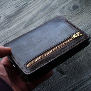 May include: A dark brown leather wallet with a gold zipper closure. The wallet has a rectangular shape and is stitched with red thread. The zipper runs diagonally across the front of the wallet.