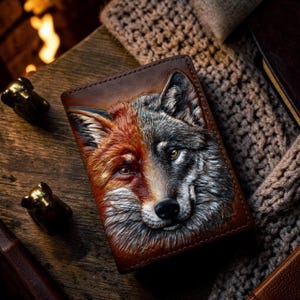 May include: A brown leather wallet featuring a detailed, embossed, and hand-painted dual-faced animal portrait. The left side depicts a reddish-orange fox, while the right side shows a grey wolf. The artwork covers the front surface of the wallet.