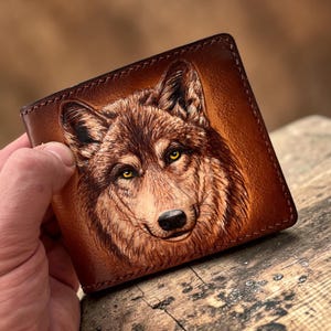 Puede incluir: Una cartera de cuero marrón con un retrato de lobo detallado, grabado y pintado a mano en la cubierta frontal. El lobo presenta texturas de pelaje realistas en tonos marrones y beige, con ojos amarillos llamativos y una nariz negra.