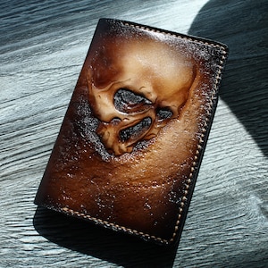 Puede incluir: Una cartera de cuero marrón con un diseño de calavera tallada. La cartera tiene un borde cosido y un aspecto desgastado.