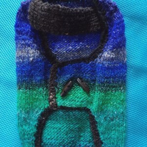 May include: A hand-knitted dog sweater in shades of blue, green, and purple. The sweater has a black collar and trim, with two wooden toggle buttons. The color gradient creates a textured appearance.