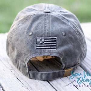 May include: A gray baseball cap with an embroidered American flag on the back. The cap has an adjustable strap with a brass buckle.
