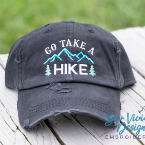 Op de afbeelding: Zwarte, verweerde baseballpet met witte borduurwerk die "Go Take A Hike" zegt met een blauwe bergketen en groene bomen.