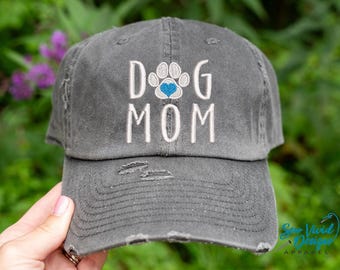 May include: A gray distressed baseball cap with the text "Dog Mom" embroidered in white with a paw print and a blue heart in the center.