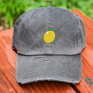 Może przedstawiać: Szara, zniszczona czapka z daszkiem z haftowanym żółtym cytryną z przodu. Cytryna ma zieloną łodygę i liść.