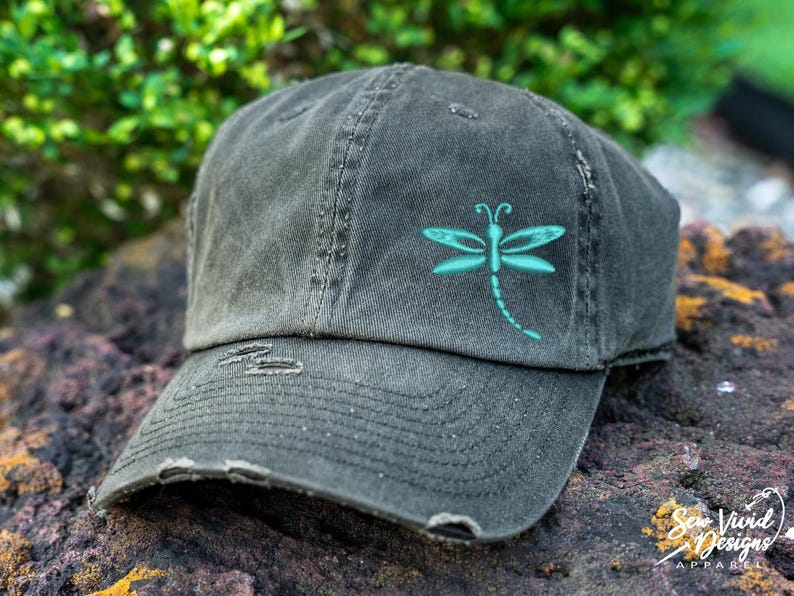 May include: A black distressed baseball cap with a teal dragonfly design embroidered on the front.