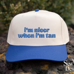 May include: A baseball cap with a white crown and a blue peak. The cap has the embroidered text "I'm nicer when I'm tan" in blue. The hat is displayed on a textured surface.