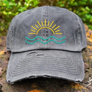 May include: Distressed grey baseball cap with an embroidered design of a yellow sun setting over teal waves. The cap has a pre-worn look with distressed details on the brim and crown. A casual accessory.
