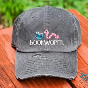 May include: A gray distressed baseball cap with an embroidered bookworm design. The bookworm is pink and has a blue book in its mouth. The text "bookworm." is embroidered in white below the bookworm.