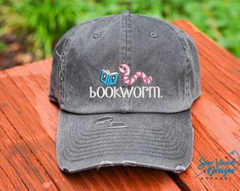 Könnte beinhalten: Eine graue, abgenutzte Baseballkappe mit einem gestickten Buchwurm-Design. Der Buchwurm ist rosa und hat ein blaues Buch im Maul. Der Text "bookworm." ist in Weiß unter dem Buchwurm gestickt.