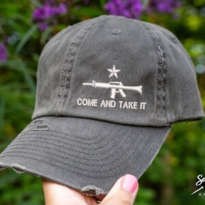 Peut inclure: Une casquette de baseball en toile grise usée avec un motif de fusil et une étoile brodés en blanc. Le texte "Come and Take It" est brodé sous le motif.