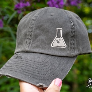 May include: A gray distressed baseball cap with a white embroidered beaker with a smiley face. The cap has a distressed look and is a great addition to any science lover's wardrobe.